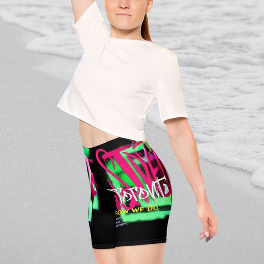 Young woman stretching on a beach wearing retro black and neon 90s biker shorts with text about Toronto and graffiti graphics | Ohhh So Swag