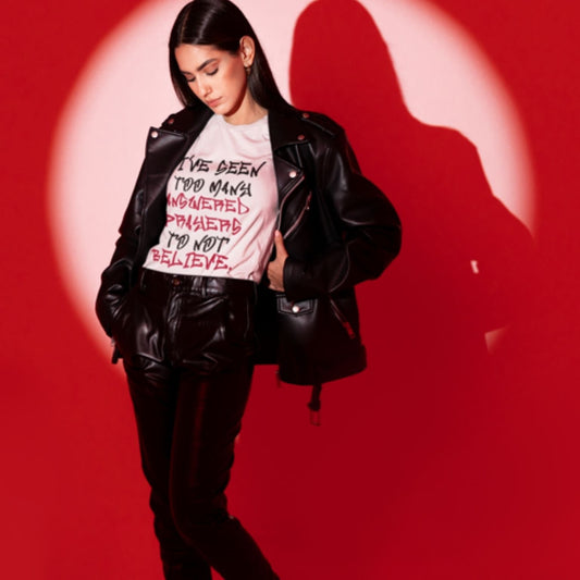 Trendy young woman wearing a black leather jacket and matching leather pants with a unisex white Christian graphic t-shirt by a red background | Ohhh So Swag