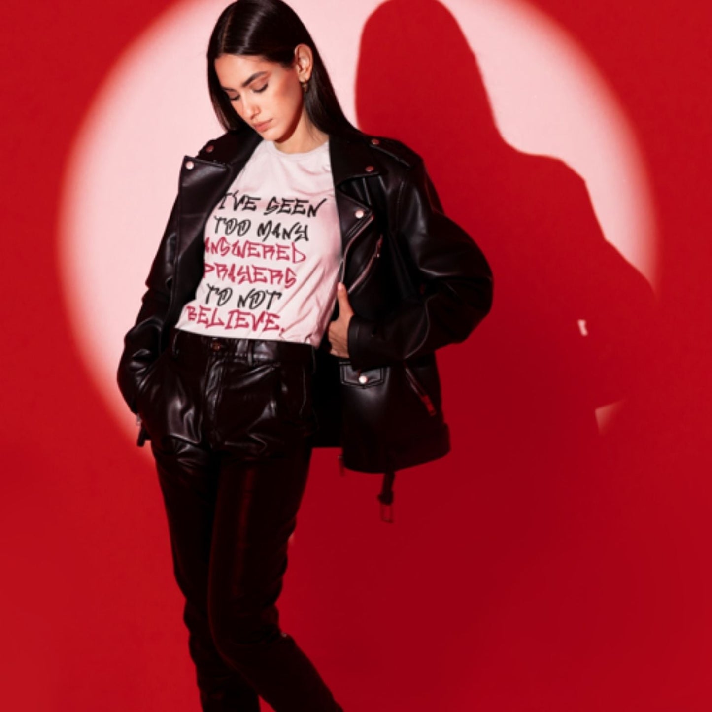 Trendy young woman wearing a black leather jacket and matching leather pants with a unisex white Christian graphic t-shirt by a red background | Ohhh So Swag