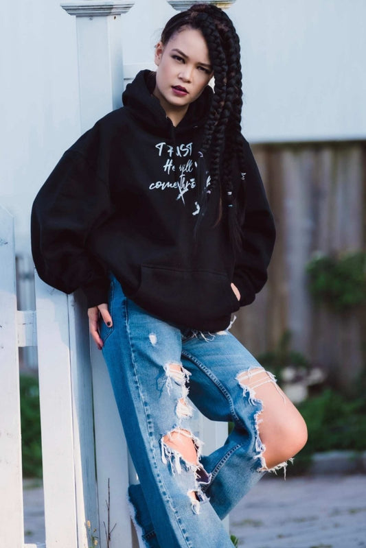 Dana Beddoe in Unisex Trust He Will Come Through Black Christian Hoodie by Ohhh So Swag and ripped jeans leaning against a white fence.