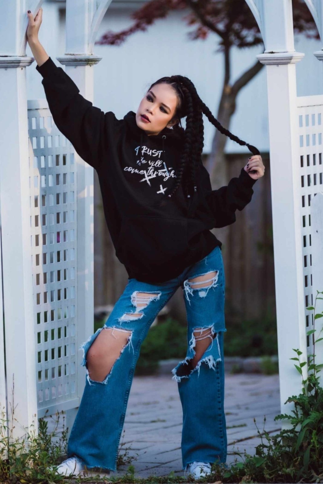 Dana Beddoe in Unisex Black Trust He Will Come Through Hoodie by Ohhh So Swag and ripped jeans against a white lattice fence.
