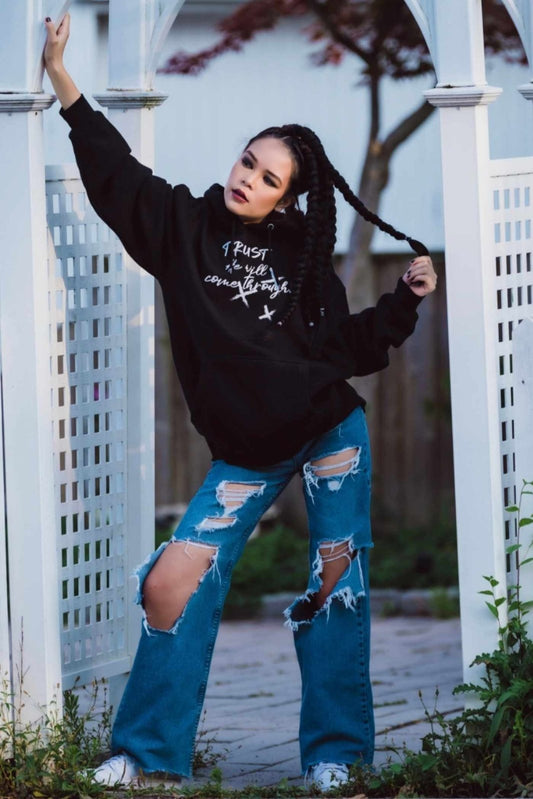 Dana Beddoe in Unisex Black Trust He Will Come Through Hoodie by Ohhh So Swag and ripped jeans against a white lattice fence.